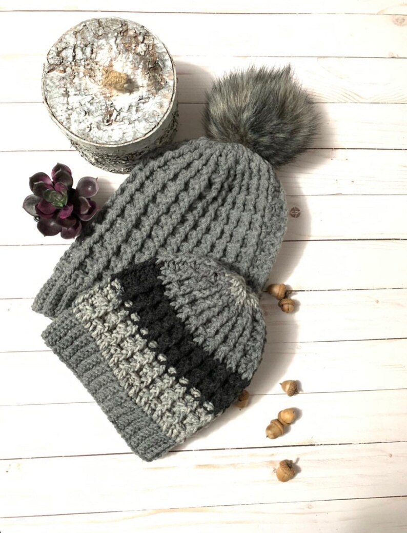May include: Two gray crocheted hats with a black stripe on the smaller hat. The larger hat has a faux fur pom pom on top. The hats are on a white wooden surface with acorns scattered around.