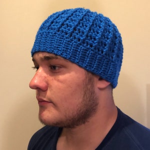 May include: A blue crocheted beanie hat with a textured pattern. The hat is worn by a person with a beard.
