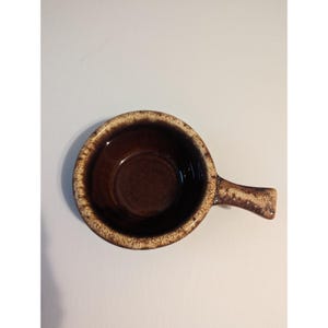 May include: A brown ceramic soup bowl with a handle. The bowl has a round shape with a dark brown interior and a lighter brown rim. The handle is small and curved, and the bowl is set against a white background.