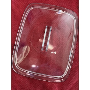 May include: A rectangular, clear glass lid featuring rounded corners and a centered, vertical, integrated handle. The lid has a raised outer rim and a smooth, transparent surface, designed to fit securely onto a corresponding container or dish.
