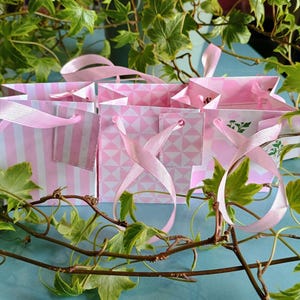 Puede incluir: Cuatro bolsas de regalo de rayas rosa y blancas con asas de cinta rosa. Las bolsas están dispuestas en fila sobre una superficie azul con hojas de hiedra verde que las rodean.