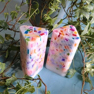 Pode incluir: Duas velas brancas retangulares com detalhes coloridos tipo confete. As velas têm um design texturizado e canelado e são encimadas por um pavio branco. Hera verde e folhas circundam as velas, sobre um fundo azul claro.