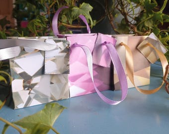 May include: Four small gift bags with ribbon handles. The bags are in various colors and patterns, including a geometric diamond pattern and solid pink. The ribbons are silver, lavender, and gold. Green foliage is in the background.