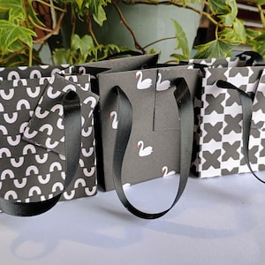 May include: Three small gift bags with black ribbon handles. The bags are made of paper and have different patterns: one with white swans on a black background, one with white and black geometric shapes, and one with white half-circles on a black background.