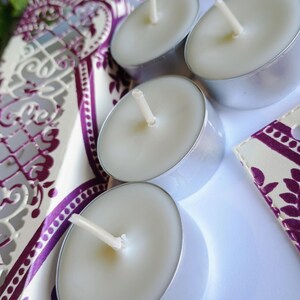 May include: Four white tea light candles with silver rims and wicks. The candles are on a white surface with a patterned fabric in the background.