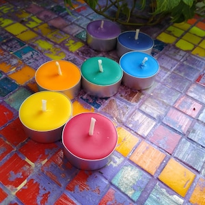 May include: A collection of seven colorful tealight candles in a variety of colors, including purple, blue, green, orange, yellow, and red. Each candle is in a silver metal container with a white wick. The candles are arranged on a mosaic tile surface.