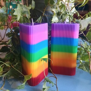 Puede incluir: Dos velas de pilar cuadradas de color arcoíris. Las velas están rayadas verticalmente con los colores del arcoíris, del rojo al morado. Las velas tienen una superficie texturizada y acanalada y una mecha blanca. Las velas se exhiben sobre una superficie azul con follaje verde.