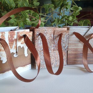 May include: Three small, decorative paper gift bags with brown satin ribbon handles. The bags have a rustic, weathered appearance with white, brown, and tan tones. The background features green foliage.