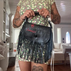 May include: A black leather handbag featuring a top handle, a long fringe trim, and a red embroidered design on the front flap. The design includes a flame symbol above a horizontal line with a decorative pattern. The bag has a structured shape.