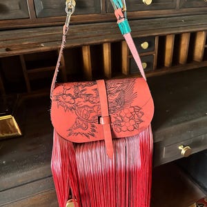 May include: A red leather shoulder bag featuring a black engraved dragon and floral design on the flap. The bag has a long red fringe, a vertical strap closure, and a multi-coloured strap with a silver chain link and a turquoise leather section.