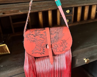 May include: A red leather shoulder bag featuring a black engraved dragon and floral design on the flap. The bag has a long red fringe, a vertical strap closure, and a multi-colored strap with a silver chain link and a turquoise leather section.