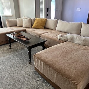 May include: A large tan sectional sofa with a chaise lounge, featuring multiple beige throw pillows and one mustard yellow pillow. A rectangular dark wood coffee table with black legs sits in front, holding a wooden tray and a small white cat.