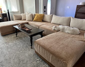 May include: A large tan sectional sofa with a chaise lounge, featuring multiple beige throw pillows and one mustard yellow pillow. A rectangular dark wood coffee table with black legs sits in front, holding a wooden tray and a small white cat.