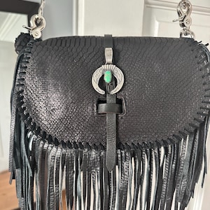 May include: A black textured leather crossbody bag featuring a long fringe trim along the bottom and sides. The flap is secured with a central black leather strap and a silver-toned horseshoe-shaped ornament set with a small oval turquoise stone.