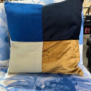 May include: Square decorative pillow with a patchwork design. The pillow features four distinct sections: blue, black, beige, and gold. The pillow is placed on a chair with a blue and white floral pattern.