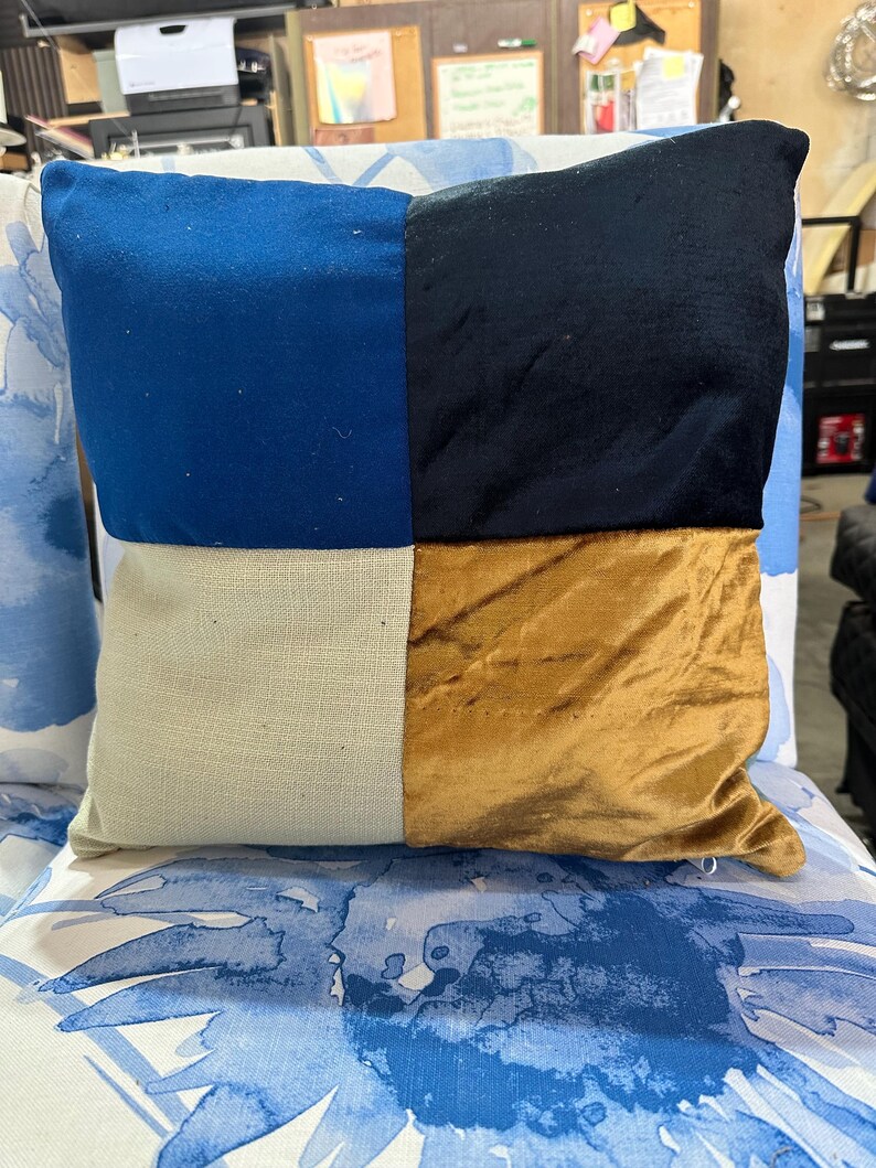 May include: Square decorative pillow with a patchwork design. The pillow features four distinct sections: blue, black, beige, and gold. The pillow is placed on a chair with a blue and white floral pattern.