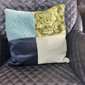 May include: A square decorative pillow with four distinct sections. One section is light blue with a quilted floral design, another is olive green with a damask pattern, a third is navy blue, and the last is a neutral beige.