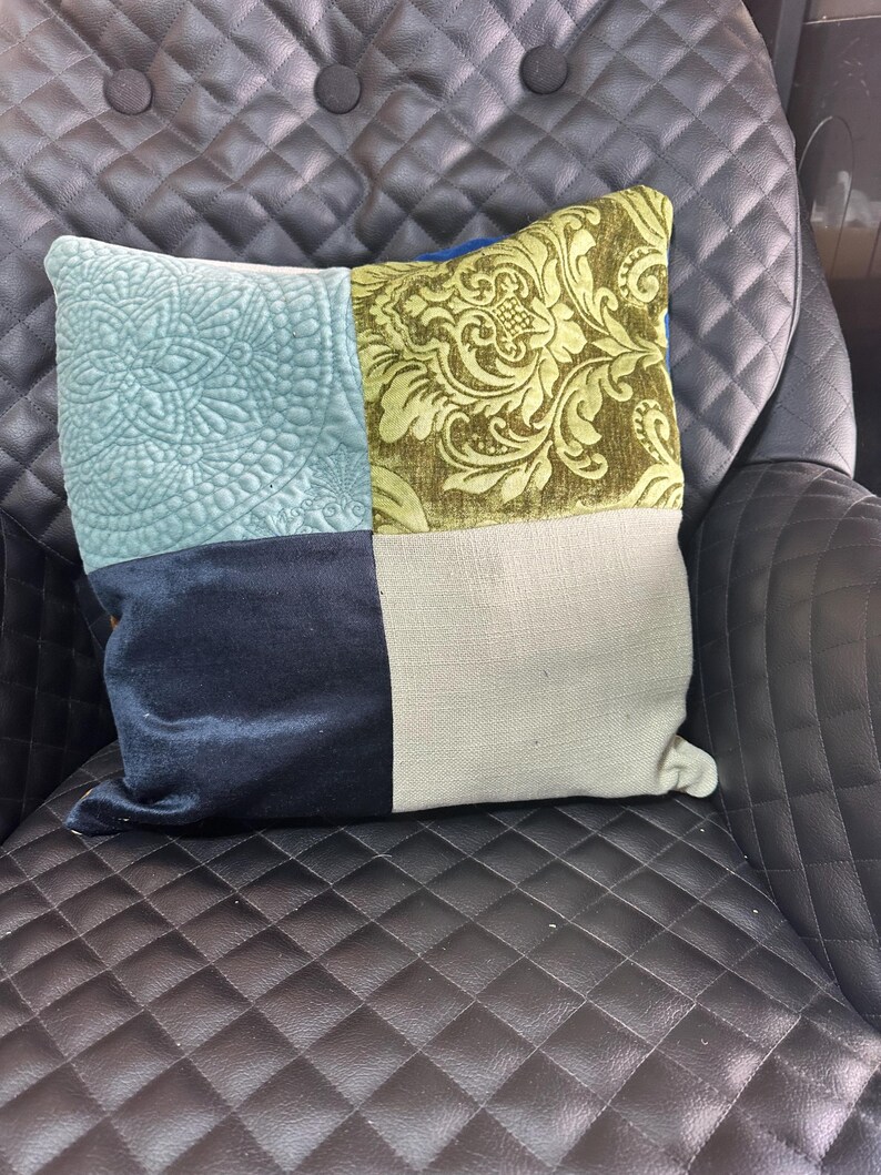 May include: A square decorative pillow with four distinct sections. One section is light blue with a quilted floral design, another is olive green with a damask pattern, a third is navy blue, and the last is a neutral beige.