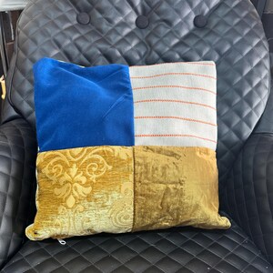 May include: A decorative square pillow with a patchwork design. The pillow features sections of blue velvet, striped fabric with orange stitching, gold damask, and gold velvet. The pillow is resting on a black leather chair.