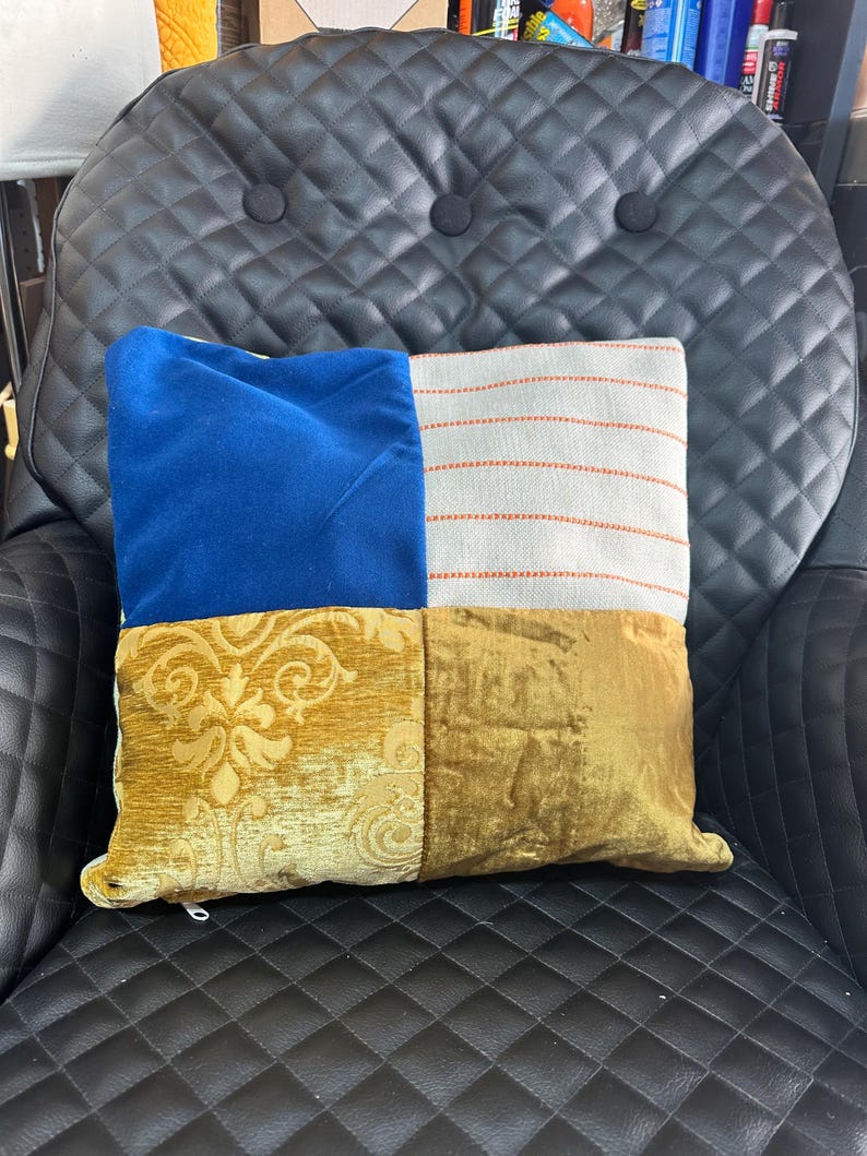 May include: A decorative square pillow with a patchwork design. The pillow features sections of blue velvet, striped fabric with orange stitching, gold damask, and gold velvet. The pillow is resting on a black leather chair.
