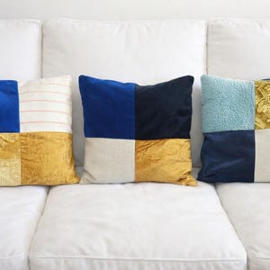 May include: Three decorative throw pillows with a patchwork design. Each pillow features a combination of colors and textures, including blue velvet, gold damask, and striped fabric. The pillows are arranged on a white sofa.