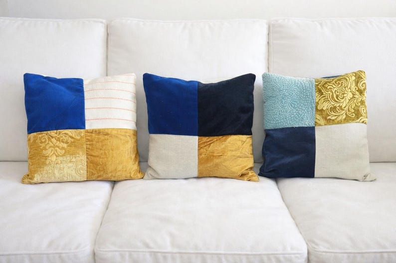 May include: Three decorative throw pillows with a patchwork design. Each pillow features a combination of colors and textures, including blue velvet, gold damask, and striped fabric. The pillows are arranged on a white sofa.