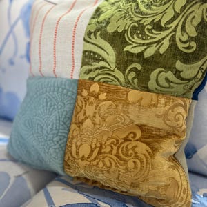 May include: A decorative patchwork pillow with four distinct fabric squares. The pillow features a light blue quilted square, a striped square with red accents, a green floral pattern, and a gold damask pattern. The pillow is resting on a blue and white patterned surface.