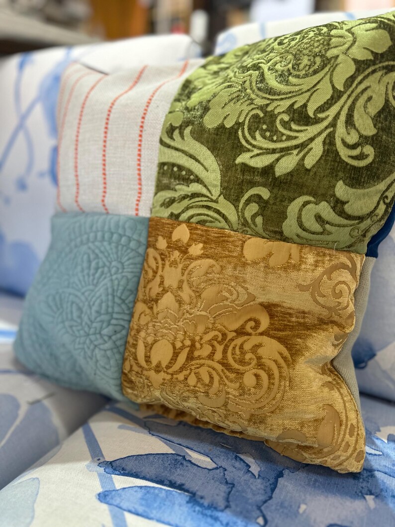 May include: A decorative patchwork pillow with four distinct fabric squares. The pillow features a light blue quilted square, a striped square with red accents, a green floral pattern, and a gold damask pattern. The pillow is resting on a blue and white patterned surface.