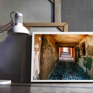 May include: A framed print of a narrow cobblestone alleyway with peeling paint and a distant view. A white desk lamp and a wooden framed canvas are in the background. The scene is set against a gray wall.