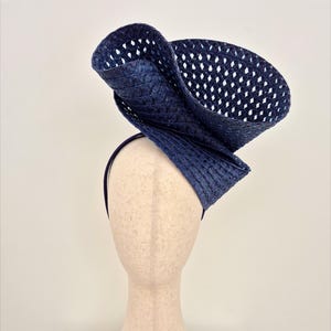 Peut inclure: Un bibi bleu fonc&eacute; dot&eacute; d'un large bord sculptural avec une texture tiss&eacute;e en treillis ouvert. La coiffe est mont&eacute;e sur un serre-t&ecirc;te fin assorti de couleur bleu fonc&eacute;, cr&eacute;ant une silhouette asym&eacute;trique et sur&eacute;lev&eacute;e avec un design pli&eacute;.