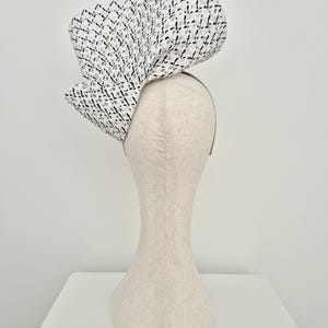 Peut inclure: Une coiffe sculpturale pr&eacute;sentant un motif en treillis tiss&eacute; blanc et noir. L'accessoire est form&eacute; de grands plis ondulants qui cr&eacute;ent une silhouette volumineuse et asym&eacute;trique. Il est mont&eacute; sur un serre-t&ecirc;te fin de couleur neutre.