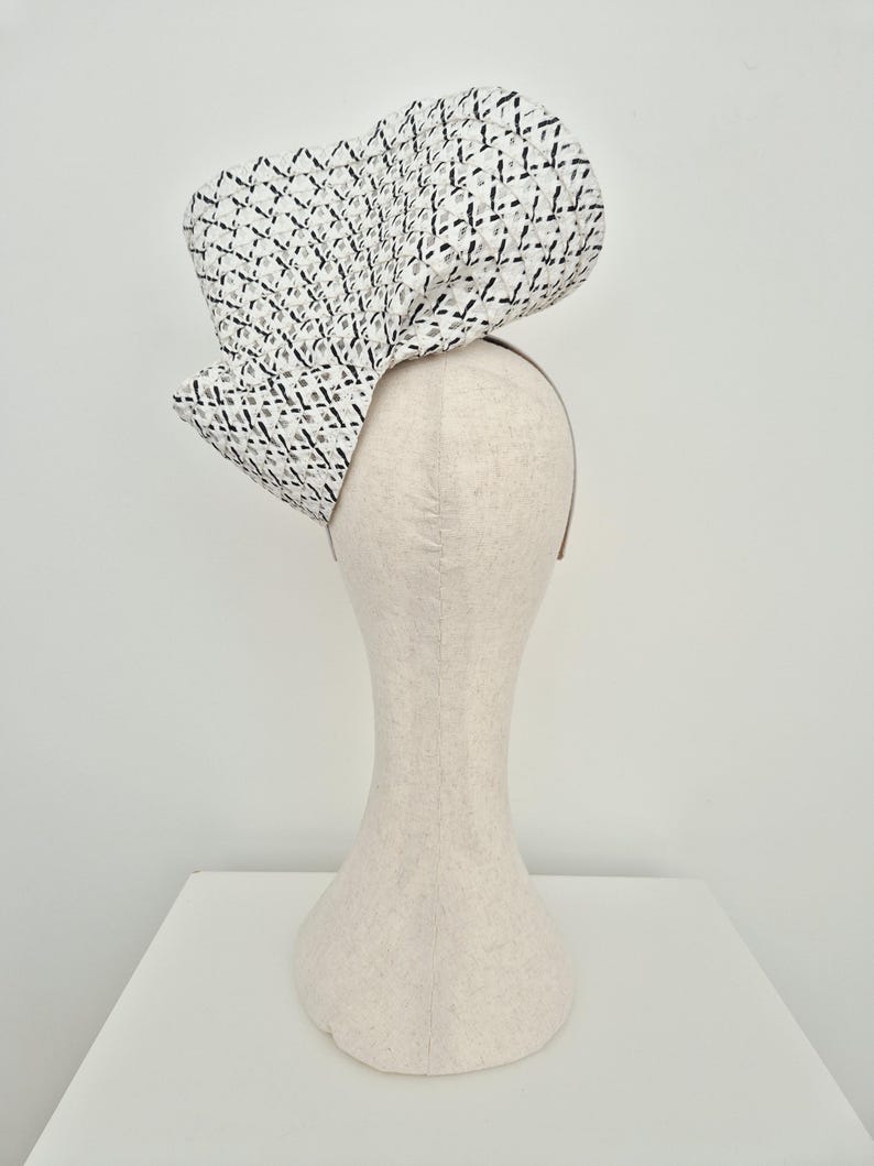 Peut inclure: Une coiffe sculpturale pr&eacute;sentant un motif en treillis tiss&eacute; blanc et noir. L'accessoire est form&eacute; de grands plis ondulants qui cr&eacute;ent une silhouette volumineuse et asym&eacute;trique. Il est mont&eacute; sur un serre-t&ecirc;te fin de couleur neutre.