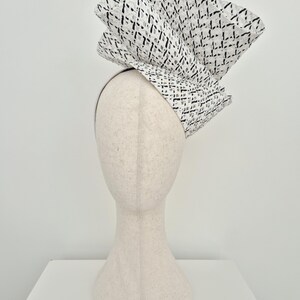 Peut inclure: Une coiffe sculpturale pr&eacute;sentant une grande structure pli&eacute;e en &eacute;ventail, faite d'un mat&eacute;riau blanc avec un motif quadrill&eacute; noir. La coiffe est mont&eacute;e sur un serre-t&ecirc;te fin de couleur neutre, cr&eacute;ant une silhouette spectaculaire et &eacute;lev&eacute;e.