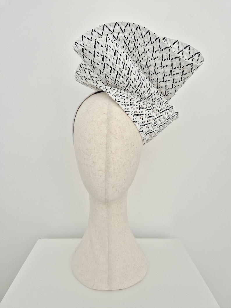 Peut inclure: Une coiffe sculpturale pr&eacute;sentant une grande structure pli&eacute;e en &eacute;ventail, faite d'un mat&eacute;riau blanc avec un motif quadrill&eacute; noir. La coiffe est mont&eacute;e sur un serre-t&ecirc;te fin de couleur neutre, cr&eacute;ant une silhouette spectaculaire et &eacute;lev&eacute;e.