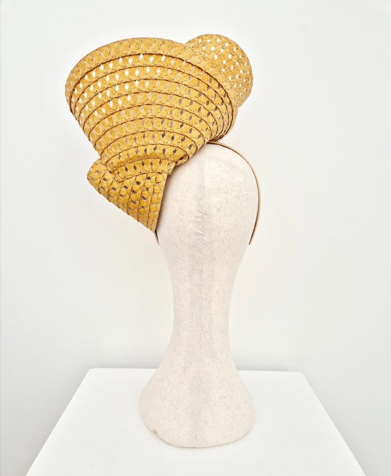 Peut inclure: Un chapeau fascinateur en paille jaune moutarde dot&eacute; d'un large bord sculpt&eacute; et tress&eacute; avec un motif ajour&eacute; en losanges. Le chapeau est mont&eacute; sur un serre-t&ecirc;te fin de couleur neutre, pr&eacute;sentant une forme et une texture structur&eacute;es et asym&eacute;triques.
