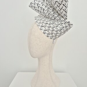 Peut inclure: Un chapeau fascinateur sculptural pr&eacute;sentant un motif en treillis tiss&eacute; blanc et noir. La coiffe est construite avec des plis architecturaux spectaculaires, repli&eacute;s et verticaux, cr&eacute;ant une silhouette asym&eacute;trique haute sur un pr&eacute;sentoir neutre.