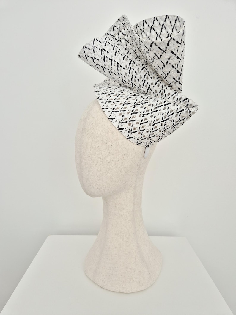 Peut inclure: Un chapeau fascinateur sculptural pr&eacute;sentant un motif en treillis tiss&eacute; blanc et noir. La coiffe est construite avec des plis architecturaux spectaculaires, repli&eacute;s et verticaux, cr&eacute;ant une silhouette asym&eacute;trique haute sur un pr&eacute;sentoir neutre.