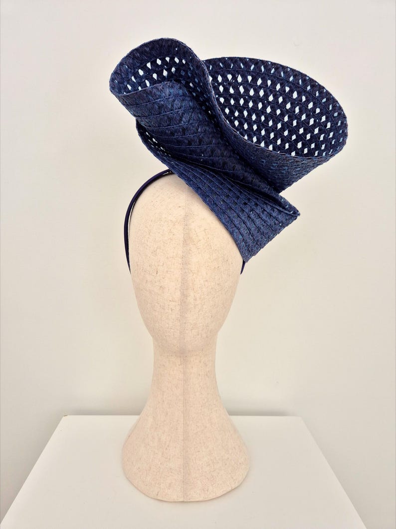 Peut inclure: Un bibi bleu fonc&eacute; dot&eacute; d'un large bord sculptural avec une texture tiss&eacute;e en treillis ouvert. La coiffe est mont&eacute;e sur un serre-t&ecirc;te fin assorti de couleur bleu fonc&eacute;, cr&eacute;ant une silhouette asym&eacute;trique et sur&eacute;lev&eacute;e avec un design pli&eacute;.