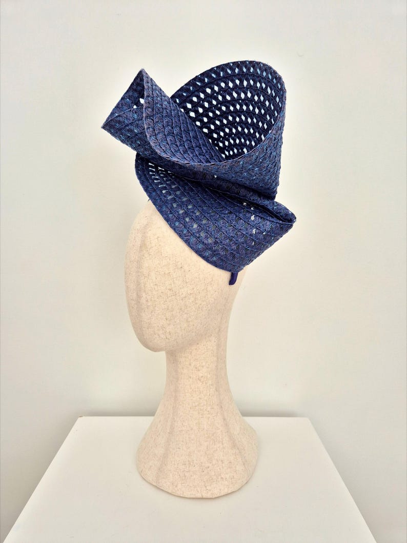 Peut inclure: Un chapeau fascinateur bleu marine profond fabriqu&eacute; en paille tress&eacute;e avec un motif en losanges ajour&eacute; distinct. La coiffe pr&eacute;sente un design haut, sculptural et asym&eacute;trique qui se courbe vers le haut et vers l'ext&eacute;rieur depuis la base.