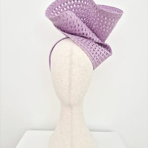 Peut inclure: Un bibi couleur lavande dot&eacute; d'un bord large, sculptural et ondul&eacute; avec une texture tiss&eacute;e ajour&eacute;e. L'accessoire est mont&eacute; sur un serre-t&ecirc;te fin assorti, cr&eacute;ant une silhouette dramatique et sur&eacute;lev&eacute;e avec un design pli&eacute; tr&egrave;s distinctif.