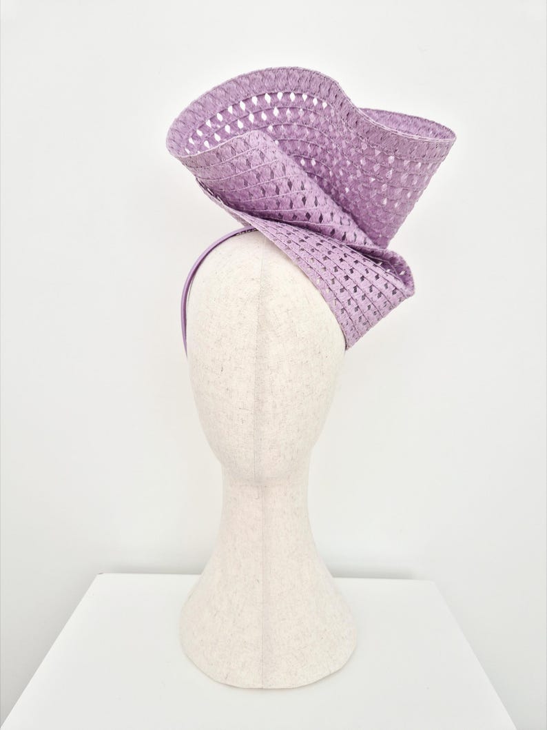 Peut inclure: Un bibi couleur lavande dot&eacute; d'un bord large, sculptural et ondul&eacute; avec une texture tiss&eacute;e ajour&eacute;e. L'accessoire est mont&eacute; sur un serre-t&ecirc;te fin assorti, cr&eacute;ant une silhouette dramatique et sur&eacute;lev&eacute;e avec un design pli&eacute; tr&egrave;s distinctif.