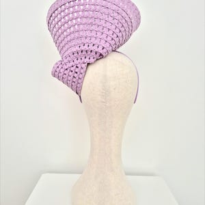 Peut inclure: Un bibi couleur lavande dot&eacute; d'un bord large et structur&eacute; avec un motif r&eacute;p&eacute;titif de d&eacute;coupes en forme de losange. Le chapeau est mont&eacute; sur un serre-t&ecirc;te fin assorti, cr&eacute;ant une silhouette sculpturale et asym&eacute;trique plac&eacute;e en hauteur.