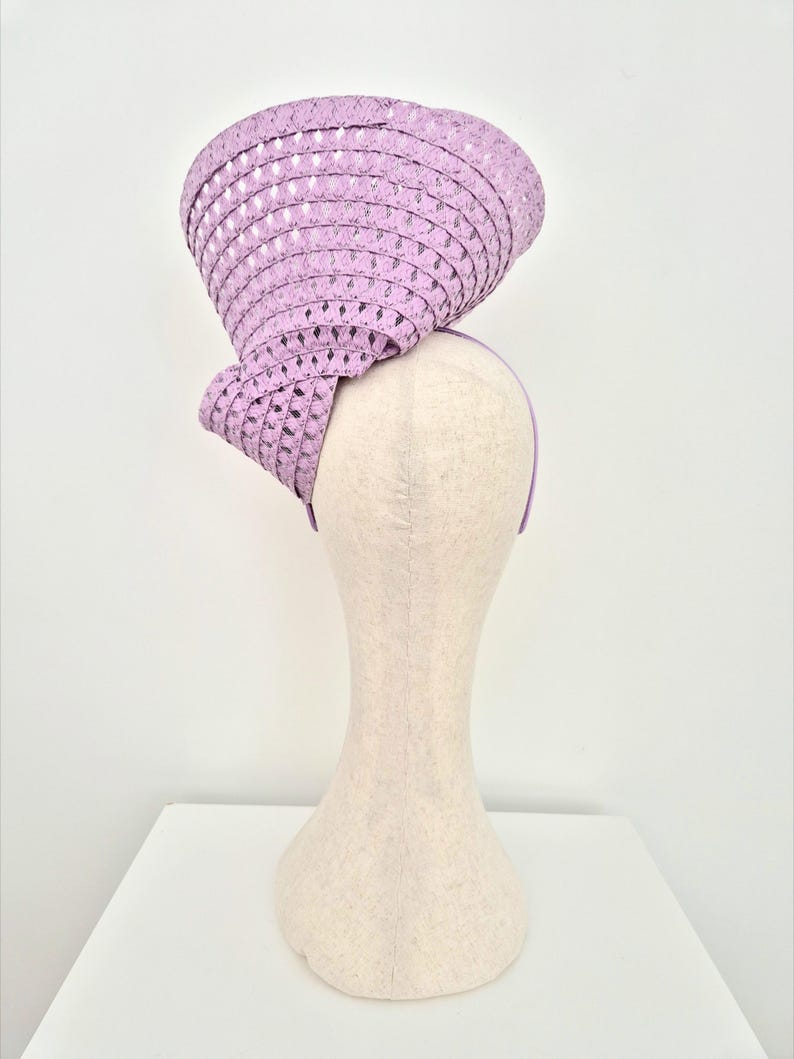 Peut inclure: Un bibi couleur lavande dot&eacute; d'un bord large et structur&eacute; avec un motif r&eacute;p&eacute;titif de d&eacute;coupes en forme de losange. Le chapeau est mont&eacute; sur un serre-t&ecirc;te fin assorti, cr&eacute;ant une silhouette sculpturale et asym&eacute;trique plac&eacute;e en hauteur.