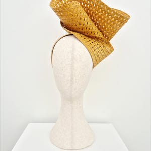 Peut inclure: Un bibi jaune moutarde dot&eacute; d'un large bord sculptural en paille tress&eacute;e avec une texture &agrave; motif losange. La coiffe est mont&eacute;e sur un serre-t&ecirc;te fin assorti, cr&eacute;ant une silhouette asym&eacute;trique et sur&eacute;lev&eacute;e qui se courbe vers le haut.