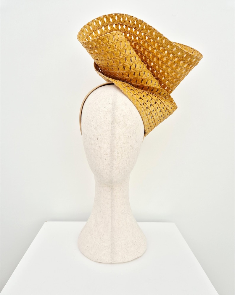 Peut inclure: Un bibi jaune moutarde dot&eacute; d'un large bord sculptural en paille tress&eacute;e avec une texture &agrave; motif losange. La coiffe est mont&eacute;e sur un serre-t&ecirc;te fin assorti, cr&eacute;ant une silhouette asym&eacute;trique et sur&eacute;lev&eacute;e qui se courbe vers le haut.