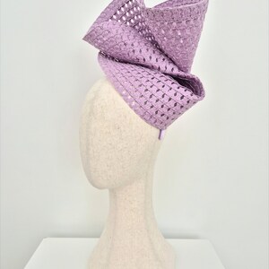 Peut inclure: Un bibi de couleur lavande pr&eacute;sentant un design sculptural et structur&eacute; avec une texture tiss&eacute;e &agrave; motif losange. La coiffe est mont&eacute;e sur un serre-t&ecirc;te fin et affiche de grands plis amples qui cr&eacute;ent une silhouette pro&eacute;minente et &eacute;lev&eacute;e.