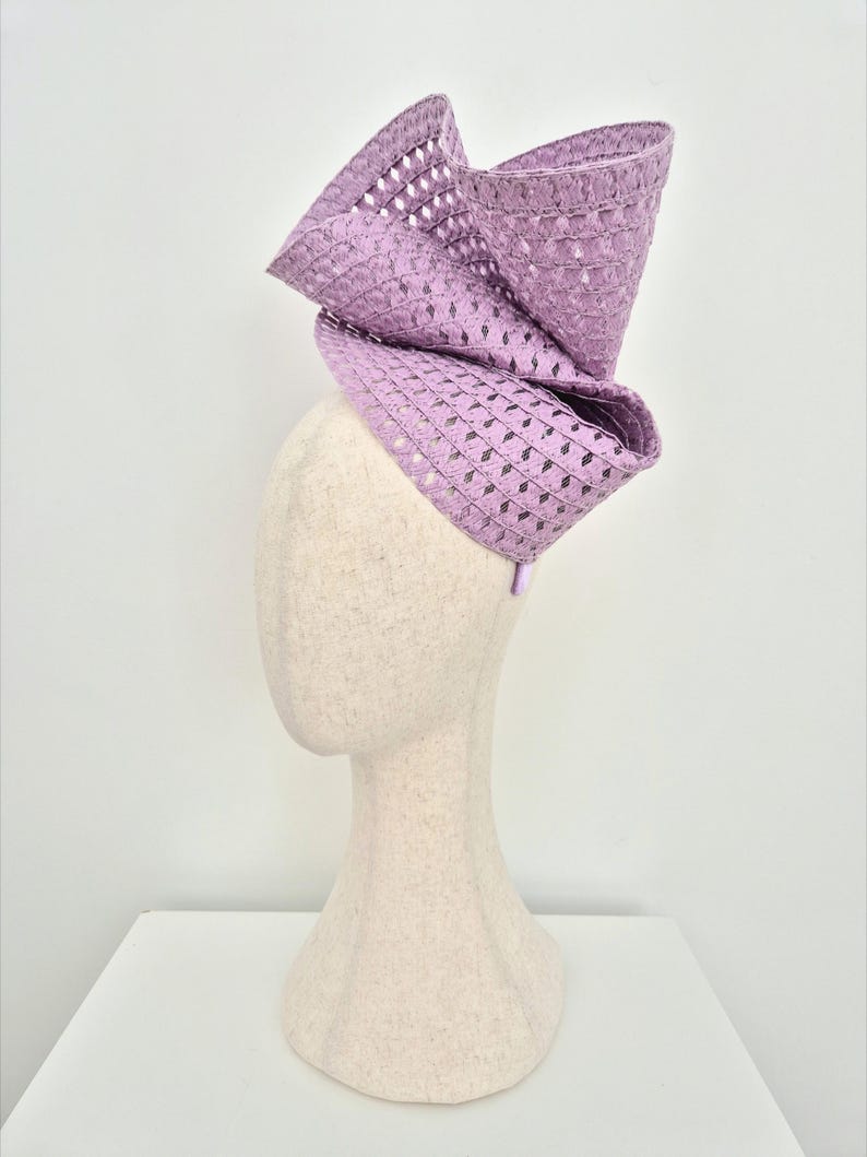 Peut inclure: Un bibi de couleur lavande pr&eacute;sentant un design sculptural et structur&eacute; avec une texture tiss&eacute;e &agrave; motif losange. La coiffe est mont&eacute;e sur un serre-t&ecirc;te fin et affiche de grands plis amples qui cr&eacute;ent une silhouette pro&eacute;minente et &eacute;lev&eacute;e.