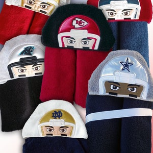 May include: Six hooded towels with embroidered football player faces. The towels are in various colors, including red, blue, black, white, and gray. The football players are wearing helmets with different team logos, including the San Francisco 49ers, New York Giants, Kansas City Chiefs, Carolina Panthers, Dallas Cowboys, and Notre Dame Fighting Irish. The towel with the Notre Dame logo has the name "PARKER" embroidered on it.