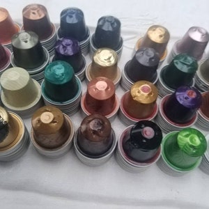 May include: A collection of colourful coffee pods, including gold, brown, green, blue, and purple, stacked in rows. The pods are made of aluminium and have a variety of designs.