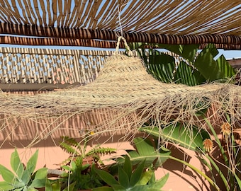 May include: A large, round, straw hat hangs from a wooden pergola covered in bamboo. The hat is made of natural fibers and has a long, flowing fringe. The hat is hanging over a lush green garden with a pink wall in the background.